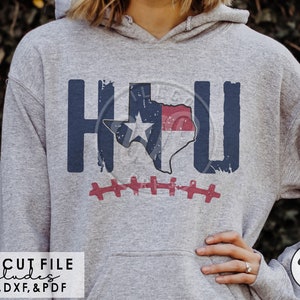May include: A heather gray hoodie featuring the letters "HOU" in navy blue, with a distressed Texas outline in the center. The Texas outline is filled with the Texas flag design. A red and white football graphic is below the letters.
