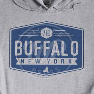 Op de afbeelding: Een grijze hoodie met een blauw en wit vintage-logo met de tekst "Buffalo New York" met een zeshoekige vorm en het nummer "716" erin.