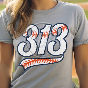 Puede incluir: Una camiseta gris con el número "313" impreso en blanco en la parte delantera. El número está bordeado de azul y tiene un diseño de puntadas de béisbol en rojo y blanco.
