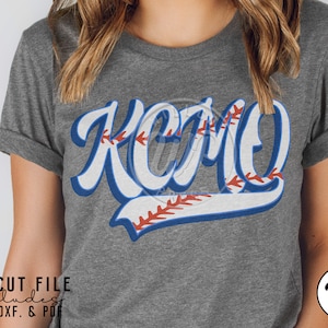 Può includere: T-shirt grigia con la scritta "KCMO" in un design a tema baseball. Le lettere sono bianche con contorni blu e dettagli di cuciture da baseball. La maglietta include formati di file SVG: PNG, DXF e PDF.