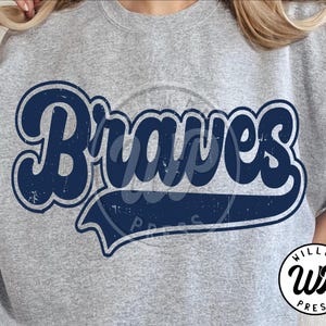 Op de afbeelding: Een grijze sweatshirt met het woord "Braves" in een marineblauwe retro-lettertype. De tekst heeft een licht versleten uitstraling, wat een vintage look geeft. Het logo van "WILLCO PRESS" is rechtsonder zichtbaar.