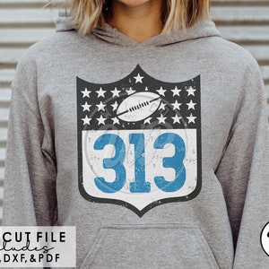 Puede incluir: Una sudadera gris con un diseño de escudo desgastado, azul y blanco con el número "313" y una pelota de fútbol. El escudo está rodeado de estrellas blancas sobre un fondo negro.