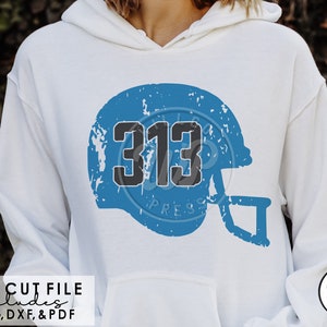 Puede incluir: Una sudadera con capucha blanca con un gráfico de casco de fútbol desgastado azul y negro con el número 313 en él. El texto "WILLCO WP PRESS" está en la esquina inferior derecha de la imagen.