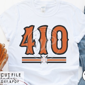 May include: A white t-shirt with a baseball-themed design featuring the number "410" in orange with black outlines. The number is centered on the shirt with two black stripes and a baseball and crossed bats graphic below.
