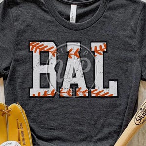 May include: A dark gray t-shirt with the word "BAL" in white letters outlined in orange. The letters are made to look like a baseball with orange stitching.
