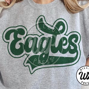 Peut inclure: Un sweat-shirt gris chiné avec le mot "Eagles" en vert délavé. Le design a un aspect rétro et vintage. Le sweat-shirt présente également un logo circulaire avec le texte "WILLCO PRESS".
