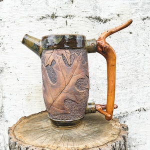May include: A brown and green ceramic pitcher with a wooden handle. The pitcher has a leaf design and is sitting on a wooden stump.