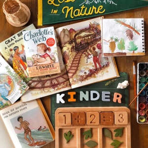 May include: A wooden counting board with numbers 1, 2, and 3, plus and equals signs. The board has five slots with leaves in each slot. The board is on a green surface with the word "kinder" in white, orange, light blue, and pink letters.