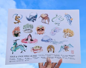May include: A watercolor print titled "Yokai & Other Magic Beings" featuring various fantastical creatures. The artwork showcases detailed illustrations of mythical beings in vibrant colors, including a snake-like figure, a crab, and a winged creature. The print is held against a bright blue sky.