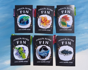 May include: Six enamel pins displayed on individual black backing cards labeled MAGICAL BEINGS PIN and www.onibaba.studio. The pins feature various illustrated creatures including a tapir, a phoenix, a character with red hair, a green lizard, a blue ray, and a fish.