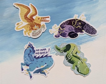 May include: Four die-cut stickers featuring illustrated creatures. One is a yellow griffin with the text 'US OUT OF EVERYWHERE', a purple platypus with lightning bolts, a blue sea horse with 'NO WAR NO BASE!', and a green salamander in water.
