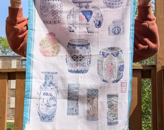 May include: A white rectangular tea towel featuring a blue border and a repeating pattern of various ceramic garden stools, vases, and jars in blue, pink, and green tones. The design includes an elephant-shaped stool and a small red stamp text.