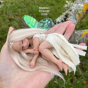 Może przedstawiać: Miniaturowa lalka wróżki z blond włosami, białą sukienką i zielonymi skrzydłami motyla. Lalka leży na dłoni, widoczny jest napis "Madelen Emmoth 2025". Wróżka to fantazyjny, ręcznie robiony przedmiot.