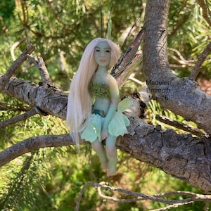 Può includere: Una bambola di fata fatta a mano con lunghi capelli biondi, un corpetto verde e una gonna verde chiaro, appollaiata su un ramo d'albero. La fata ha ali delicate ed è ambientata su uno sfondo verde sfocato. Il testo sull'immagine recita: Madelen Emmoth Maj-26.