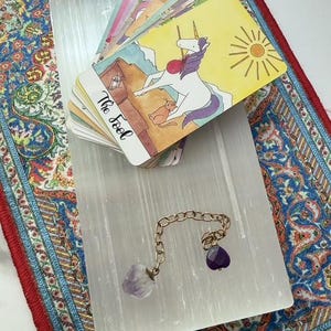 May include: A deck of tarot cards featuring a unicorn illustration on the top card labeled The Fool, resting on a rectangular white selenite slab. A gold-toned metal chain with a raw amethyst crystal and a small purple heart-shaped charm is attached.