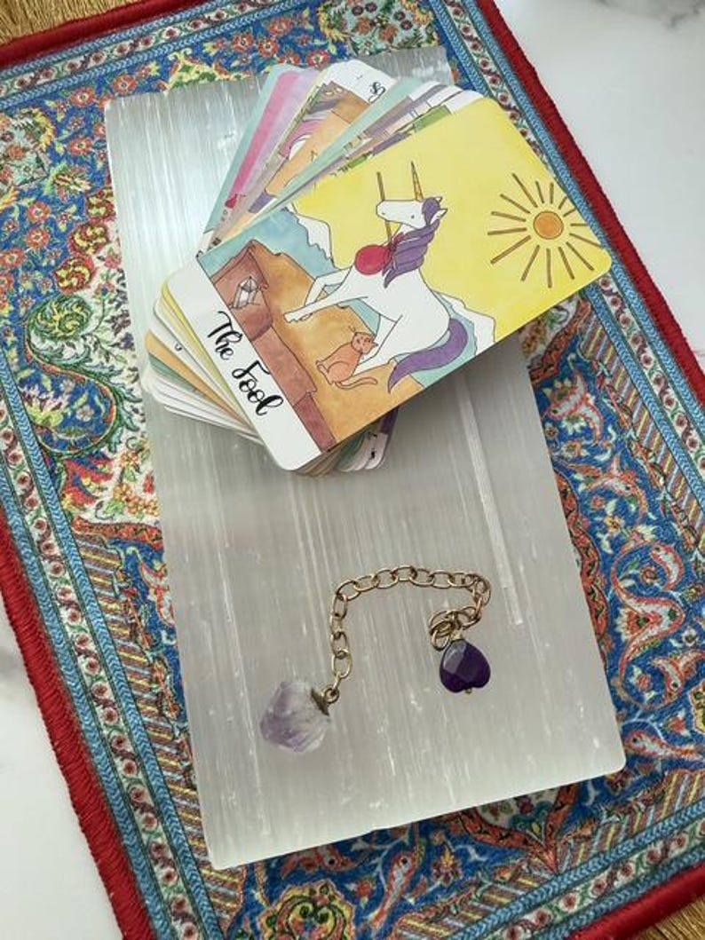 May include: A deck of tarot cards featuring a unicorn illustration on the top card labeled The Fool, resting on a rectangular white selenite slab. A gold-toned metal chain with a raw amethyst crystal and a small purple heart-shaped charm is attached.