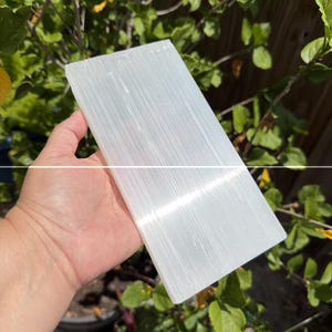 May include: A rectangular slab of white selenite crystal with a fibrous, translucent texture and a smooth, flat surface. The stone displays natural vertical striations and a pearlescent sheen that reflects light across its elongated, flat form.