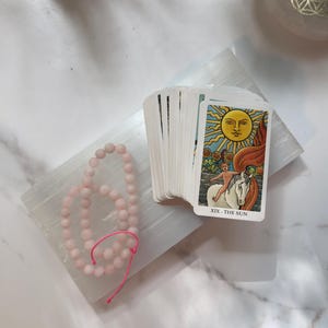 May include: A deck of tarot cards featuring the XIX The Sun card on top, showing a yellow sun, a child on a white horse, and sunflowers. Beside the deck is a strand of light pink matte beads on a bright pink string, all resting on a white slab.