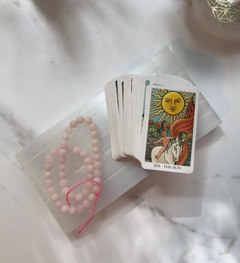 May include: A deck of tarot cards featuring the XIX The Sun card on top, showing a yellow sun, a child on a white horse, and sunflowers. Beside the deck is a strand of light pink matte beads on a bright pink string, all resting on a white slab.