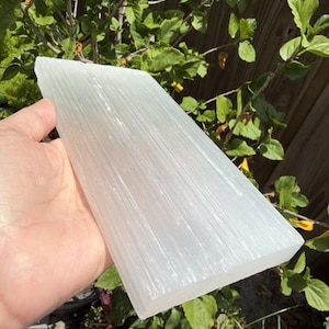 May include: A rectangular slab of white selenite crystal with a translucent appearance and a fibrous, linear texture running along its length. The surface of the stone displays a natural, shimmering luster and a smooth, flat finish on its edges.