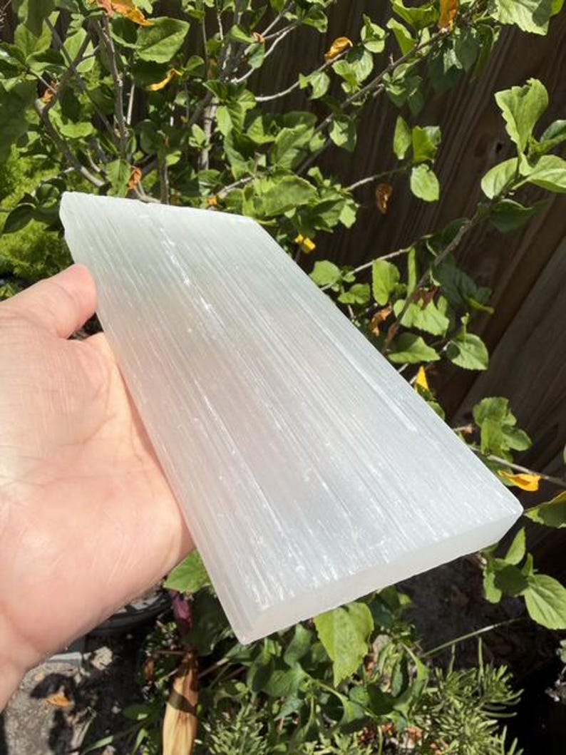 May include: A rectangular slab of white selenite crystal with a translucent appearance and a fibrous, linear texture running along its length. The surface of the stone displays a natural, shimmering luster and a smooth, flat finish on its edges.
