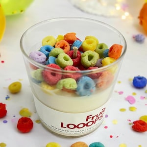 May include: A clear glass jar candle with a white wax base and a colorful layer of cereal loops on top. The jar has a white label with the text "Fruit Loops" in red and "StudioMarLeif Etsy" in black. The candle is surrounded by colorful confetti.