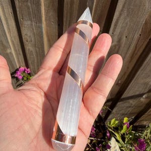 May include: A clear, elongated crystal wand with a pointed tip, wrapped with copper-colored metal. The crystal is held in a hand, with a wooden fence and greenery in the background. The wand is approximately 6 inches long.