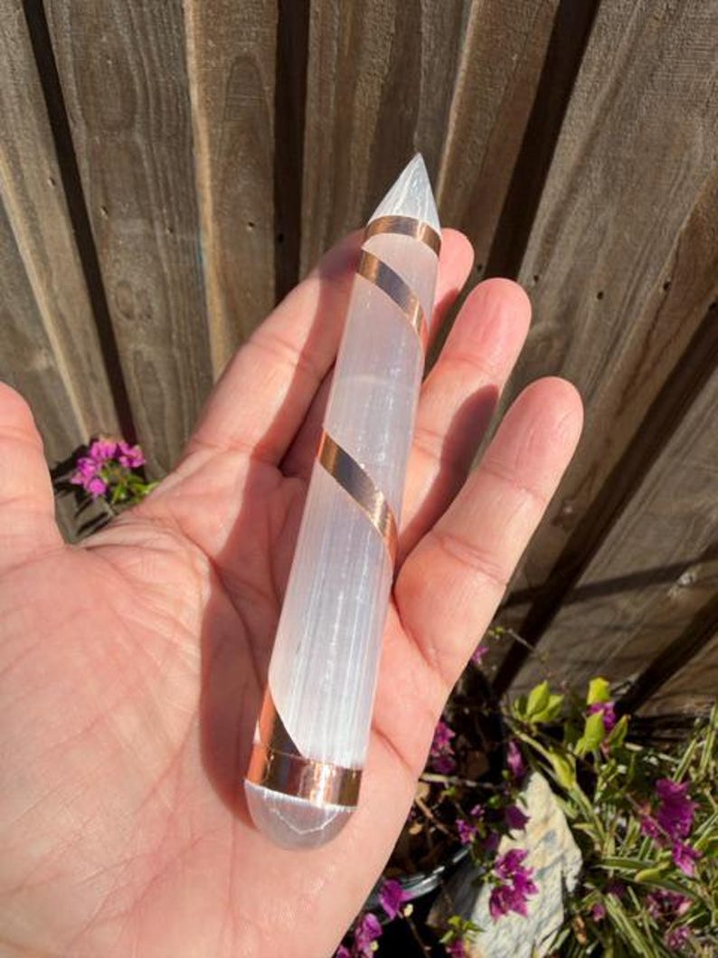 May include: A clear, elongated crystal wand with a pointed tip, wrapped with copper-colored metal. The crystal is held in a hand, with a wooden fence and greenery in the background. The wand is approximately 6 inches long.