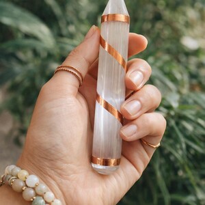 May include: A clear, elongated crystal point with copper-colored bands spiraling around it. The crystal is held in a hand, with rings on the fingers and a beaded bracelet on the wrist. The background is blurred green foliage.