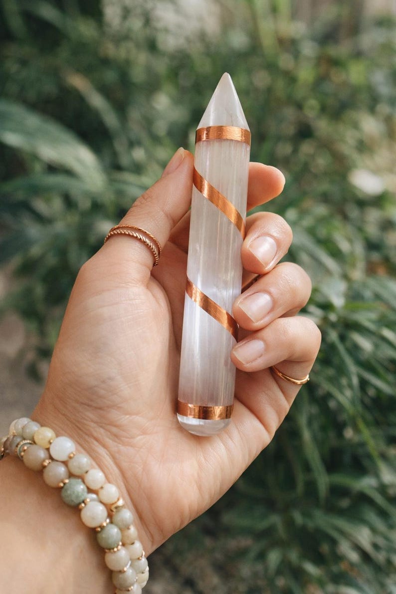 May include: A clear, elongated crystal point with copper-colored bands spiraling around it. The crystal is held in a hand, with rings on the fingers and a beaded bracelet on the wrist. The background is blurred green foliage.