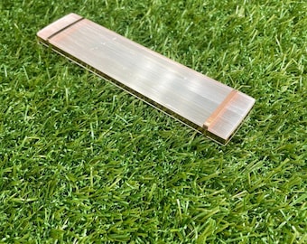 May include: A rectangular, translucent selenite crystal wand with a copper-colored band. The wand is lying on a bed of green grass. The crystal is approximately 6 inches long.