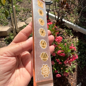 May include: A rectangular selenite crystal wand with gold-colored chakra symbols etched on its surface. The wand has a clear, translucent appearance with a copper-colored band at the top and bottom.