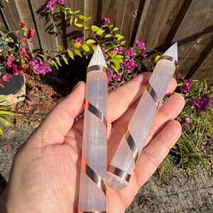 May include: Two selenite wands with copper-colored spiral accents. The wands are held in a hand, with a wooden fence and flowering plants in the background. The wands have a clear, crystalline appearance.