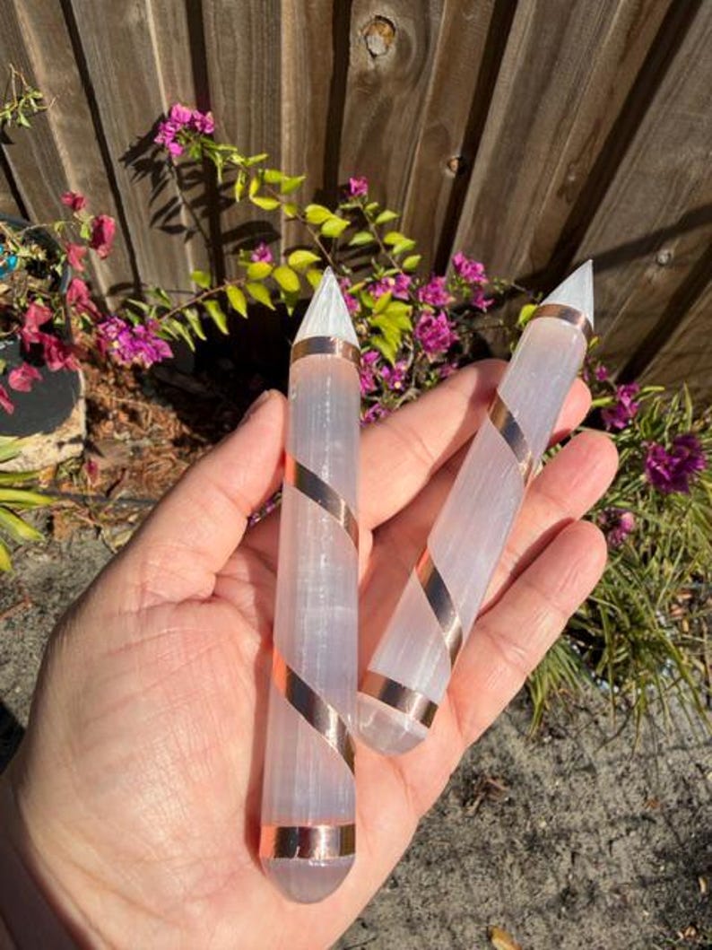 May include: Two selenite wands with copper-colored spiral accents. The wands are held in a hand, with a wooden fence and flowering plants in the background. The wands have a clear, crystalline appearance.