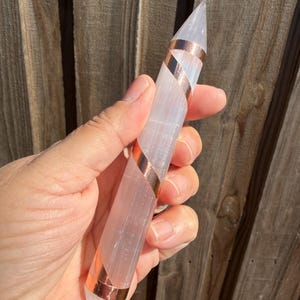 May include: A clear, elongated crystal wand with a pointed tip, wrapped with copper bands. The crystal is held in a hand, with a wooden fence in the background. The wand is approximately 6 inches long.