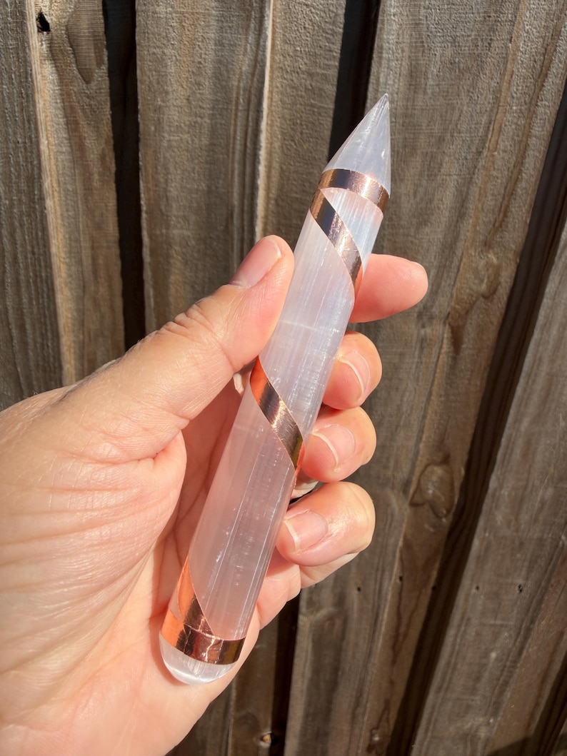 May include: A clear, elongated crystal wand with a pointed tip, wrapped with copper bands. The crystal is held in a hand, with a wooden fence in the background. The wand is approximately 6 inches long.