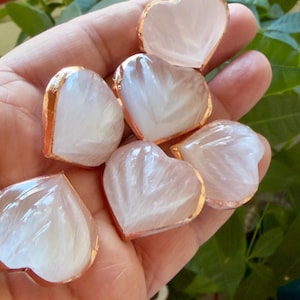 Puede incluir: Una colección de piedras en forma de corazón, translúcidas, rosas y blancas, con un borde dorado. Las piedras se sostienen en una mano, con follaje verde en el fondo. Los corazones tienen un patrón interno plumoso.