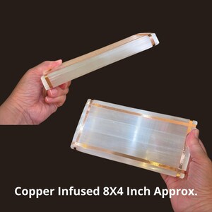 May include: A rectangular selenite slab featuring copper-colored metal strips wrapped around the edges. The white, translucent stone has a fibrous texture. The text Copper Infused 8X4 Inch Approx. is visible at the bottom of the image.