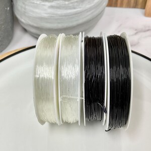 May include: Four spools of elastic cord, two clear and two black, are arranged on a white plate. The spools are all the same size and have a white plastic base.