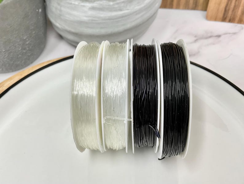 May include: Four spools of elastic cord, two clear and two black, are arranged on a white plate. The spools are all the same size and have a white plastic base.