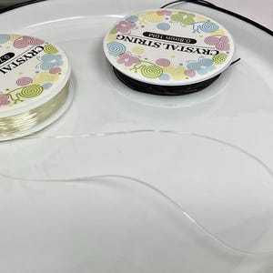 May include: Two spools of clear elastic string for jewelry making. The spools are labeled "Crystal String" and have a colorful floral design. The string is 0.7mm thick and comes in 10m lengths.