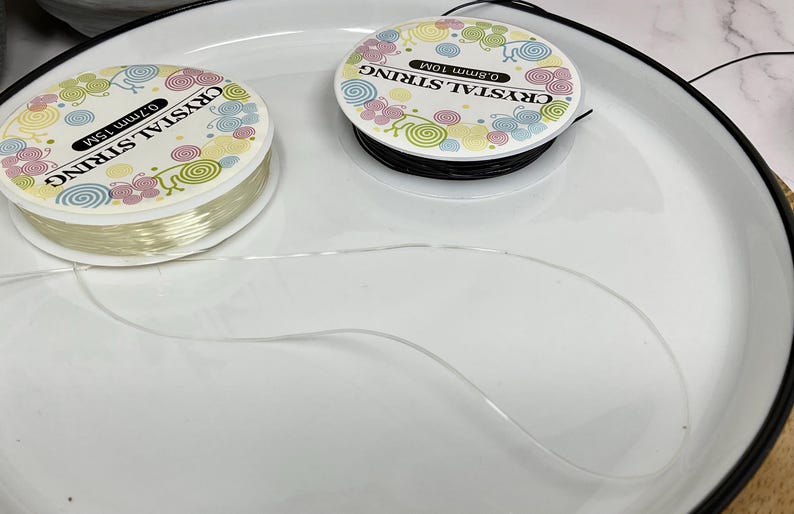 May include: Two spools of clear elastic string for jewelry making. The spools are labeled "Crystal String" and have a colorful floral design. The string is 0.7mm thick and comes in 10m lengths.