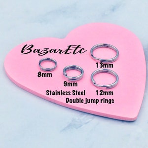 May include: Four stainless steel double jump rings in different sizes: 8mm, 9mm, 12mm, and 13mm. The rings are displayed on a pink heart-shaped surface.
