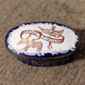 May include: Oval trinket box with a white top featuring a blue border and a decorative edge. The top has a design of two birds and a red ribbon with the words "AMANS FIDELLE". The box has a blue base and a gold-colored metal rim.