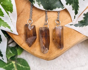 May include: Three brown smoky quartz crystal pendants on silver chains. The crystals are all pointed and have a natural, raw look. The pendants are displayed on a wooden surface.