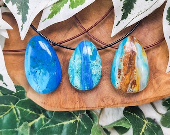 May include: Three polished gemstone pendants in shades of blue and brown, each suspended on a black or brown cord. The gemstones have unique patterns and textures. The pendants are displayed on a wooden surface, surrounded by green and white leaves.
