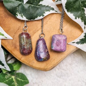 Può includere: Tre ciondoli in pietra preziosa su catene d'argento. I ciondoli sono rosso scuro, viola e rosa chiaro. I ciondoli sono tutti di forme e dimensioni diverse. I ciondoli sono su una superficie di legno con foglie verdi sullo sfondo.