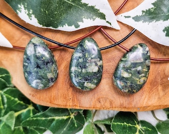 May include: Three green and white speckled stone pendants on brown leather cords. The pendants are teardrop shaped and are displayed on a wooden surface.