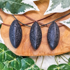 Peut inclure: Trois pendentifs noirs de forme ovale sur un collier en cordon noir. Les pendentifs ont une surface rugueuse et texturée. Le collier est présenté sur une surface en bois avec des feuilles vertes.