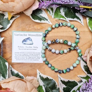 Pode incluir: Duas pulseiras de variscite wavellite verde e branca sobre uma superfície de madeira com um cartão que diz "Variscite Wavellite Meditação - Conhecimento - Cura "Minha alma e meu espírito estão curados." A variscite wavellite é uma pedra poderosa para ajudar a deixar de lado o julgamento. Traz amor incondicional e compreensão em todas as situações. Ajuda a olhar para o "panorama geral" antes de tomar uma decisão. A pedra ajuda a fornecer informações espirituais para a conclusão das tarefas. © Crystal Cave"